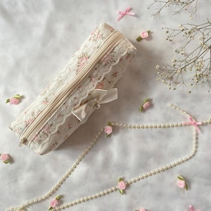 K&ouml;nnte beinhalten: Eine wei&szlig;e Stofftasche mit Blumenmuster, R&auml;nder aus Spitze und einem Perlen-Rei&szlig;verschluss. Die Tasche ist mit einem wei&szlig;en Satinband gebunden und von rosa Satinrosen und einer Perlenkette umgeben.