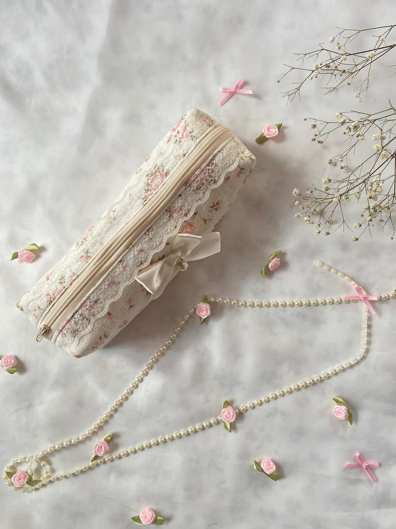 K&ouml;nnte beinhalten: Eine wei&szlig;e Stofftasche mit Blumenmuster, R&auml;nder aus Spitze und einem Perlen-Rei&szlig;verschluss. Die Tasche ist mit einem wei&szlig;en Satinband gebunden und von rosa Satinrosen und einer Perlenkette umgeben.