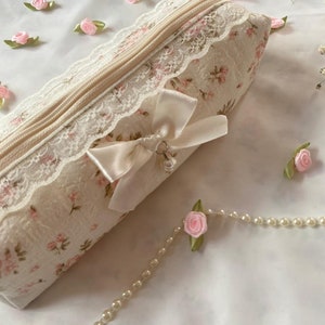 K&ouml;nnte beinhalten: Eine wei&szlig;e Rei&szlig;verschlusstasche mit Blumenmuster und Spitzenbesatz. Die Tasche hat ein wei&szlig;es Satinband, das mit einer Schleife und einem Metallanh&auml;nger gebunden ist. Die Tasche ist von kleinen rosa Stoffblumen und einer Perlenkette umgeben.