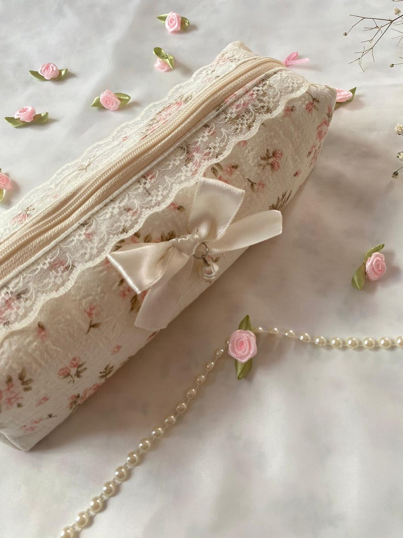K&ouml;nnte beinhalten: Eine wei&szlig;e Rei&szlig;verschlusstasche mit Blumenmuster und Spitzenbesatz. Die Tasche hat ein wei&szlig;es Satinband, das mit einer Schleife und einem Metallanh&auml;nger gebunden ist. Die Tasche ist von kleinen rosa Stoffblumen und einer Perlenkette umgeben.