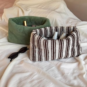 May include: Two soft, rectangular fabric storage baskets. One is solid sage green, and the other features vertical brown and cream stripes. Both contain small cosmetic bottles. A pair of black oval sunglasses rests nearby on the white fabric.