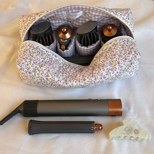 Könnte beinhalten: Eine Kosmetiktasche mit floralem Muster und Vichy-Futter, die Haarstyling-Tools enthält. Ein Haartrockner, ein Lockenstab und eine weiße Haarspange mit floralem Design sind ebenfalls sichtbar. Die Tasche trägt den Text "cozy things".