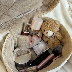 May include: A beige, zippered cosmetic bag labeled "cozy things" filled with makeup, a small teddy bear, and hair clips. The bag contains a variety of beauty products, including a compact, mascara, and lipstick. The teddy bear has a brown bow.