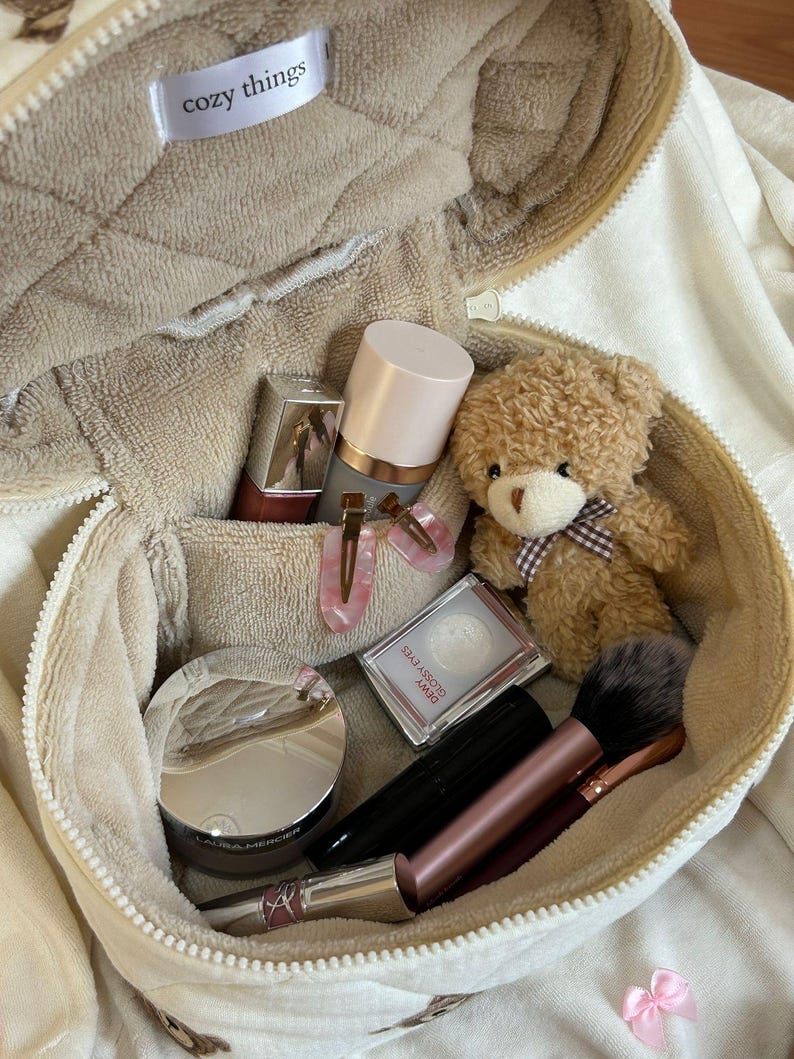May include: A beige, zippered cosmetic bag labeled "cozy things" filled with makeup, a small teddy bear, and hair clips. The bag contains a variety of beauty products, including a compact, mascara, and lipstick. The teddy bear has a brown bow.