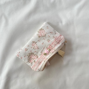 May include: A small white zippered pouch with a pink floral pattern and a pink rose embellishment. The pouch is lying on a white surface.