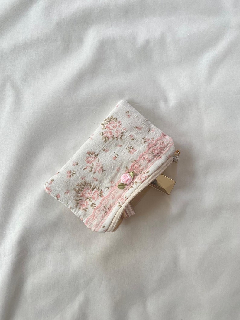 May include: A small white zippered pouch with a pink floral pattern and a pink rose embellishment. The pouch is lying on a white surface.