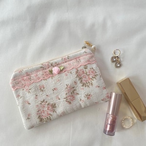 May include: A small white zippered pouch with a pink floral pattern and lace trim. The pouch has a pink bow and a gold zipper pull with a pearl charm. The pouch is lying on a white surface with a gold lipstick and a gold ring.