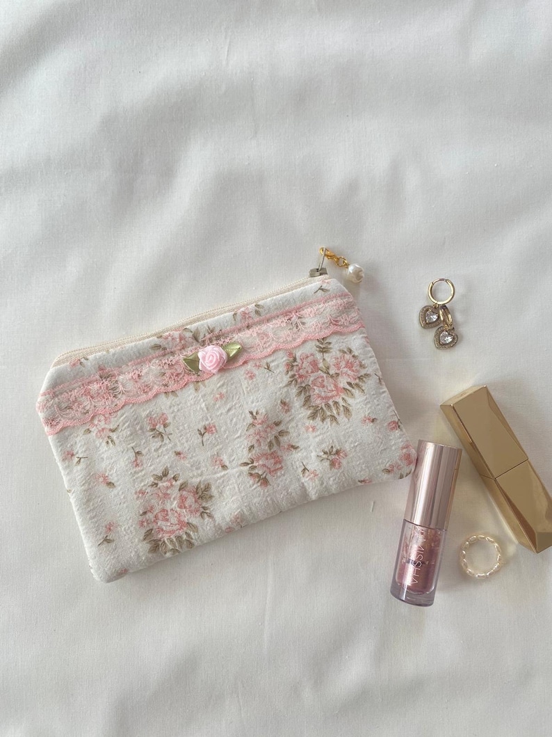 May include: A small white zippered pouch with a pink floral pattern and lace trim. The pouch has a pink bow and a gold zipper pull with a pearl charm. The pouch is lying on a white surface with a gold lipstick and a gold ring.