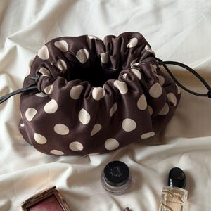 Può includere: Un beauty case in raso marrone con coulisse e grandi pois bianchi. La borsa è parzialmente aperta, rivelando un interno scuro. Articoli per il trucco, tra cui un compatto, un vasetto e un flacone di profumo, sono disposti intorno alla borsa su un tessuto color crema.