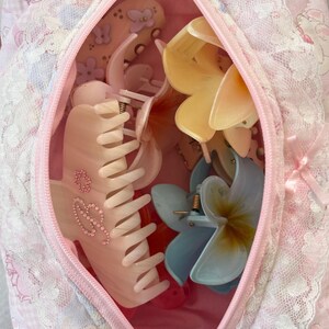 May include: A pink zippered cosmetic bag filled with various hair clips. The clips are in the shape of flowers and claw clips in shades of pink, yellow, and blue. The bag is decorated with lace and a floral pattern.