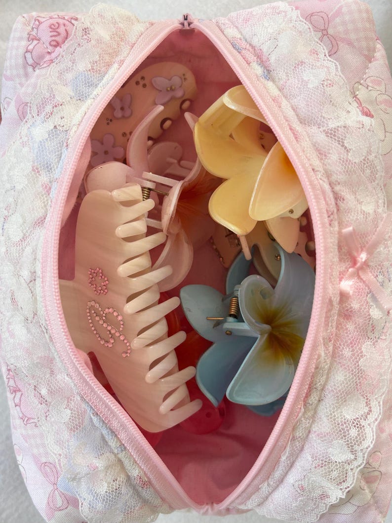 May include: A pink zippered cosmetic bag filled with various hair clips. The clips are in the shape of flowers and claw clips in shades of pink, yellow, and blue. The bag is decorated with lace and a floral pattern.