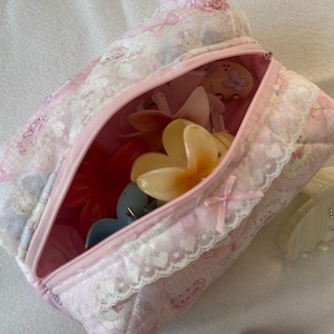 May include: A pink and white patterned cosmetic bag with a zipper, filled with various hair clips. The bag has a lace trim and a small bow. The bag is on a white surface.