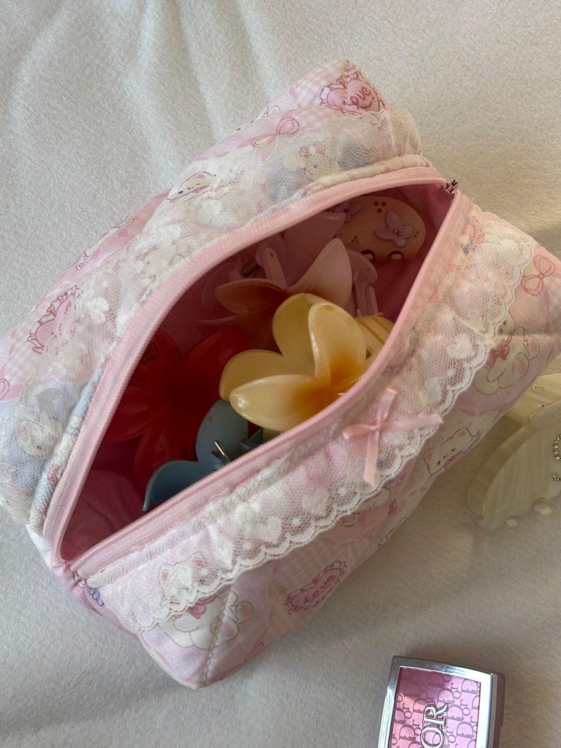 May include: A pink and white patterned cosmetic bag with a zipper, filled with various hair clips. The bag has a lace trim and a small bow. The bag is on a white surface.
