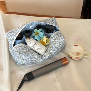 Könnte beinhalten: Eine Kosmetiktasche mit floralem Muster mit verschiedenen Haarstyling-Werkzeugen, darunter ein Haartrockner und Aufsätze. Eine Flasche Haarprodukt und eine Tasse Kaffee sind ebenfalls vorhanden.