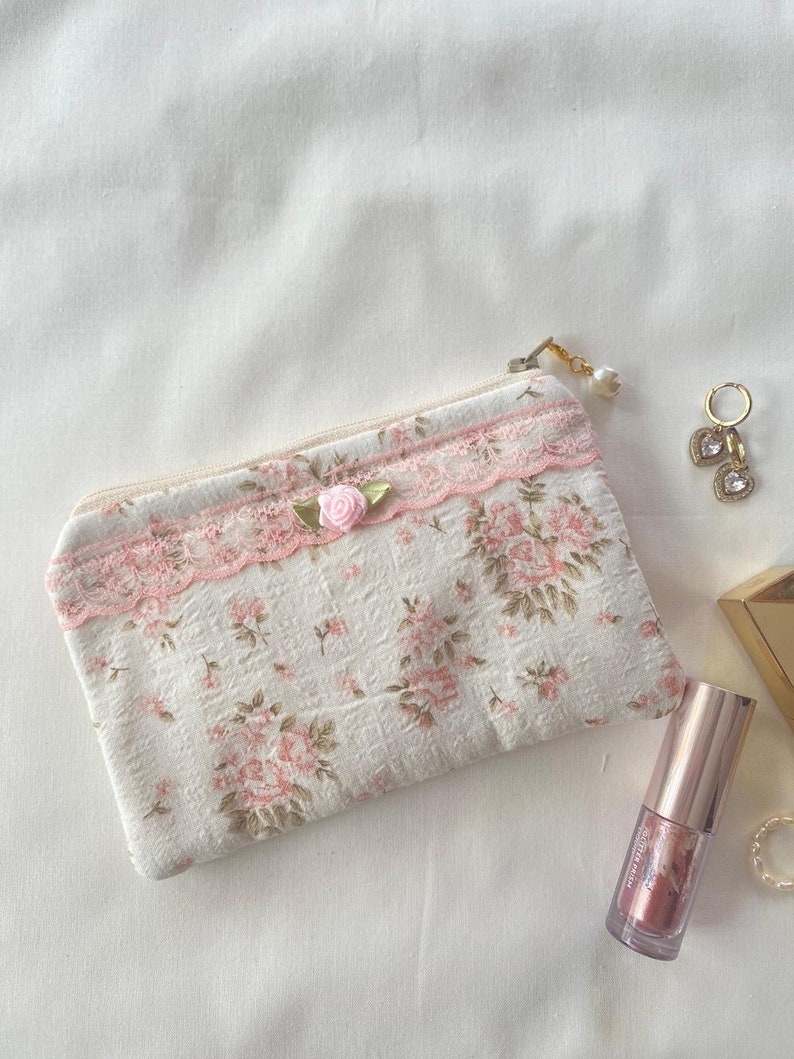 May include: A small white zippered pouch with a pink floral pattern and lace trim. A pink rose embellishment is attached to the zipper pull. The pouch is lying on a white surface.