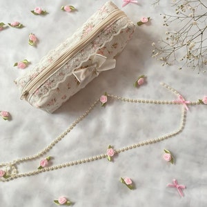 K&ouml;nnte beinhalten: Eine wei&szlig;e Stofftasche mit Blumenmuster und Spitzenbesatz. Die Tasche hat einen Rei&szlig;verschluss und eine wei&szlig;e Schleife aus Satin. Die Tasche ist von rosa Stoffblumen und einer Perlenkette umgeben.