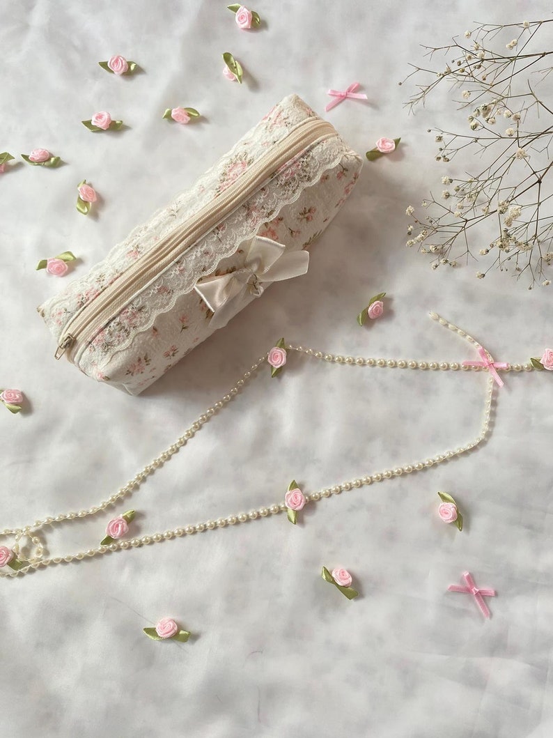 K&ouml;nnte beinhalten: Eine wei&szlig;e Stofftasche mit Blumenmuster und Spitzenbesatz. Die Tasche hat einen Rei&szlig;verschluss und eine wei&szlig;e Schleife aus Satin. Die Tasche ist von rosa Stoffblumen und einer Perlenkette umgeben.