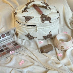 May include: A quilted, off-white makeup bag with a repeating pattern of brown teddy bears. The bag is open, revealing a black zipper and a pink lip gloss. A makeup palette, a lipstick, and a mug are also present.