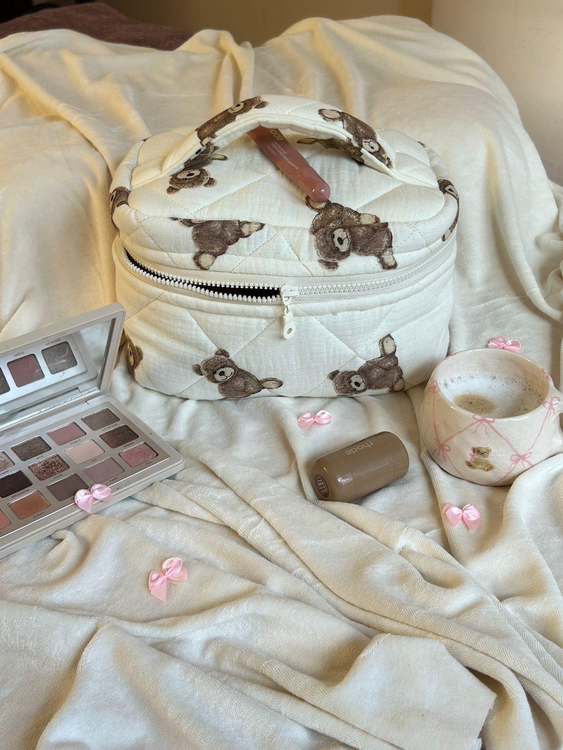 May include: A quilted, off-white makeup bag with a repeating pattern of brown teddy bears. The bag is open, revealing a black zipper and a pink lip gloss. A makeup palette, a lipstick, and a mug are also present.