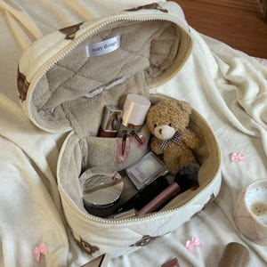 May include: Open, quilted cosmetic bag with a teddy bear pattern. The bag is filled with makeup products, a small teddy bear, and a makeup brush. The interior is lined with a soft, beige material. The text "cozy things" is visible inside the bag.