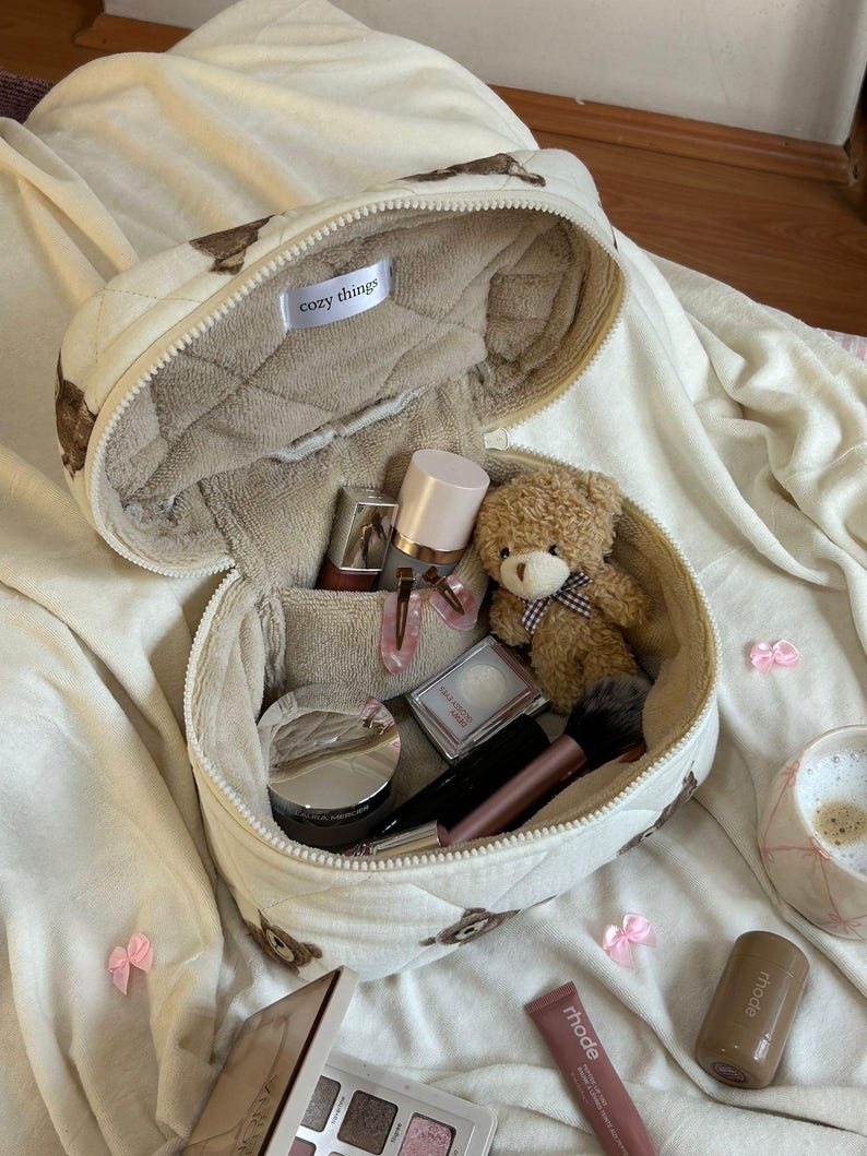 May include: Open, quilted cosmetic bag with a teddy bear pattern. The bag is filled with makeup products, a small teddy bear, and a makeup brush. The interior is lined with a soft, beige material. The text "cozy things" is visible inside the bag.