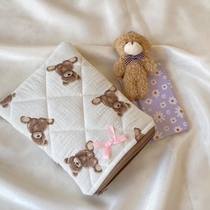 Könnte beinhalten: Eine weiße gesteppte Reißverschlusstasche mit einem braunen Bärenmuster und einem rosa Band. Ein kleiner brauner Teddybär mit einer karierten Schleife liegt neben der Tasche auf einem weißen Stoffhintergrund.