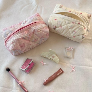 May include: Two quilted cosmetic bags, one pink and one cream, with matching makeup accessories. The pink bag features a pattern with characters and lace trim. Accessories include a blush compact, lip gloss, a makeup brush, and a hair clip, all adorned with pink bows.