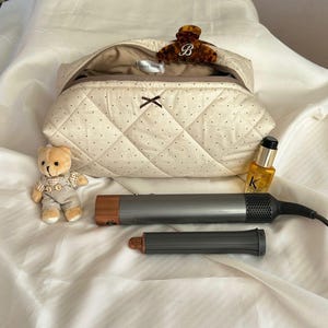 Puede incluir: Un neceser acolchado beige con lunares negros, un lazo marrón y una pinza para el pelo carey con una inicial "B". También hay un osito de peluche, herramientas para peinar el cabello y una botella de aceite capilar.