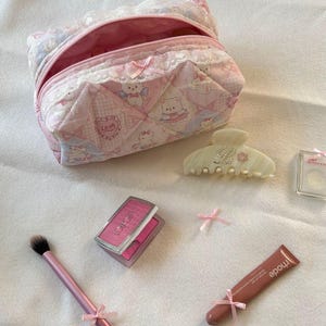 May include: A pink quilted cosmetic bag with a teddy bear pattern, open to reveal a pink interior. Makeup items include a blush compact, brush, hair clip, lip tint, and small pink bows. The items are arranged on a white surface.