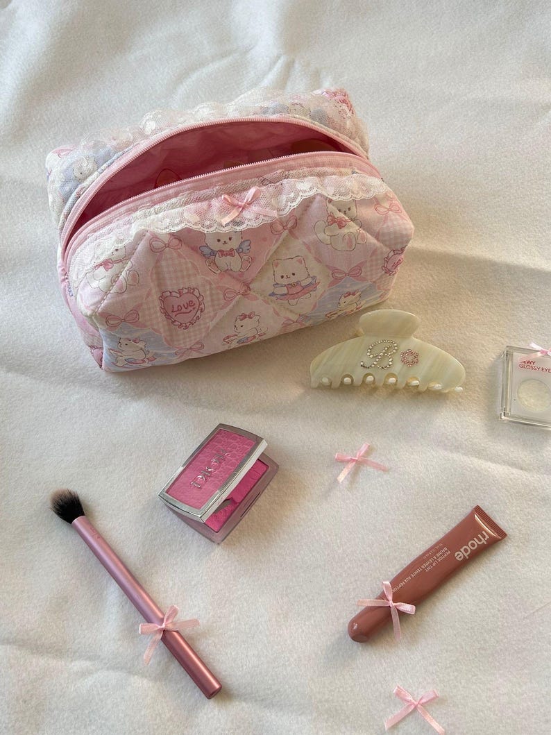 May include: A pink quilted cosmetic bag with a teddy bear pattern, open to reveal a pink interior. Makeup items include a blush compact, brush, hair clip, lip tint, and small pink bows. The items are arranged on a white surface.