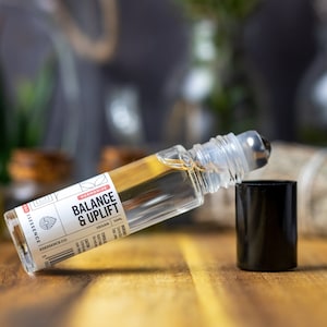 May include: A clear glass roller bottle with a black cap and a silver roller ball. The bottle is labelled "Harmony Essence Co. Balance & Uplift Vegan 10ml".
