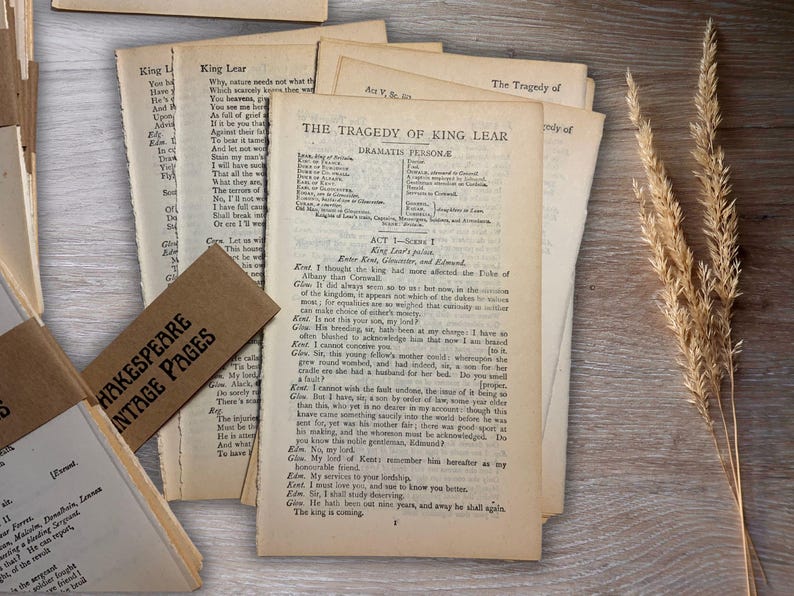 May include: Vintage pages from Shakespeare's "King Lear" are displayed on a wooden surface. The pages are aged with visible text, and a brown tag reads "Shakespeare Vintage Pages." A sprig of dried wheat adds a rustic touch.