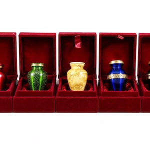 May include: Five small, decorative urns in a row, each displayed in a red velvet box. The urns are in various colors, including red, green, yellow, and blue, with gold-colored accents. The boxes are open, revealing the urns.