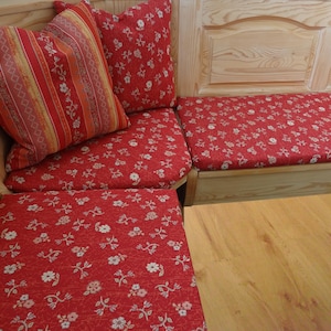 Könnte beinhalten: Eine Eckbank aus hellem Holz mit roten Sitz- und Rückenpolstern mit weißem Blumenmuster. Ein quadratisches Zierkissen zeigt ein rot-orange-gelbes Streifenmuster, während ein weiteres quadratisches Kissen zu den floralen Sitzpolstern passt.