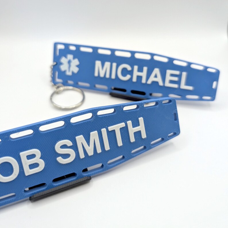 Puede incluir: Dos etiquetas de identificaci&oacute;n m&eacute;dica de pl&aacute;stico azul con texto blanco. La etiqueta superior dice "MICHAEL" y la etiqueta inferior dice "BOB SMITH". Ambas etiquetas tienen un peque&ntilde;o s&iacute;mbolo de estrella de la vida.