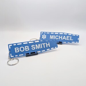 Puede incluir: Dos llaveros de metal azul con letras blancas. El llavero superior tiene un s&iacute;mbolo m&eacute;dico y el nombre "MICHAEL". El llavero inferior tiene el nombre "BOB SMITH".