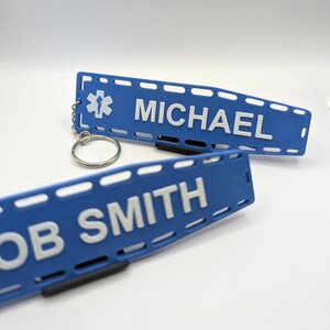 Puede incluir: Dos etiquetas de identificaci&oacute;n m&eacute;dica de pl&aacute;stico azul con texto blanco. La etiqueta superior tiene un s&iacute;mbolo m&eacute;dico blanco y el nombre "MICHAEL". La etiqueta inferior tiene el nombre "BOB SMITH".