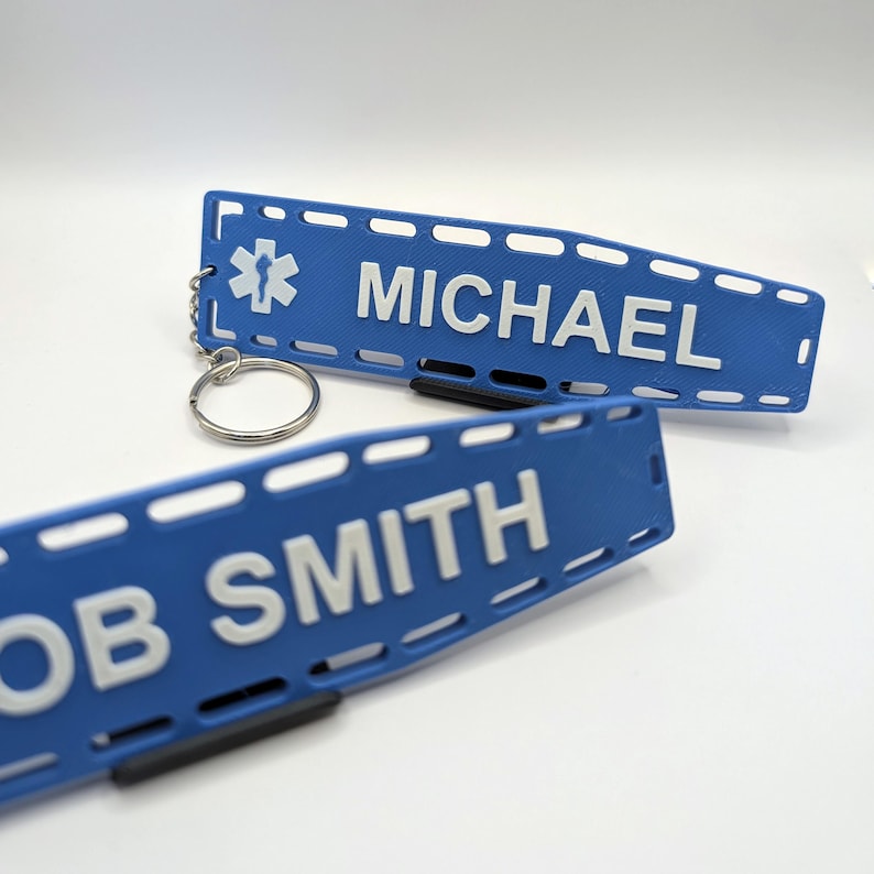 Puede incluir: Dos etiquetas de identificaci&oacute;n m&eacute;dica de pl&aacute;stico azul con texto blanco. La etiqueta superior tiene un s&iacute;mbolo m&eacute;dico blanco y el nombre "MICHAEL". La etiqueta inferior tiene el nombre "BOB SMITH".