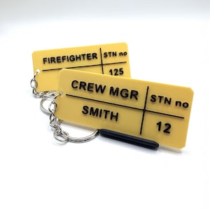 Puede incluir: Dos llaveros de plástico amarillo con texto negro. El llavero superior dice "FIREFIGHTER" y "STN no 125". El llavero inferior dice "CREW MGR SMITH" y "STN no 12".