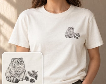 May include: An off-white short-sleeve t-shirt featuring a small, detailed black ink-style illustration on the left chest. The graphic depicts a fluffy Pallas's cat sitting next to a small pine branch with two pine cones.