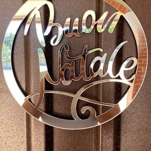 Könnte beinhalten: Ein silberner runder Anhänger mit den Worten "Buon Natale" in einer dekorativen Schreibschrift ausgeschnitten.