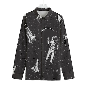Puede incluir: Una camisa negra de manga larga con botones y estampado de temática espacial. El estampado presenta estrellas blancas, un astronauta blanco y naves espaciales blancas.