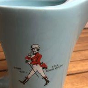 May include: A light blue ceramic pitcher with a handle and a spout. The pitcher features a graphic of a man in a red coat and the text "BORN 1820 - STILL GOING STRONG". The pitcher is on a wooden surface.