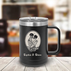 May include: Black stainless steel travel mug with a black handle. The mug has a white design of two skeletons kissing inside a circle with a crescent moon and stars. The text "Sophia & Steve" is printed below the design.