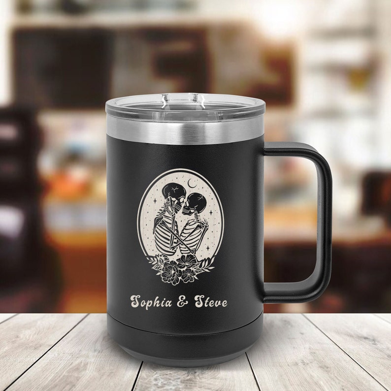 May include: Black stainless steel travel mug with a black handle. The mug has a white design of two skeletons kissing inside a circle with a crescent moon and stars. The text "Sophia & Steve" is printed below the design.