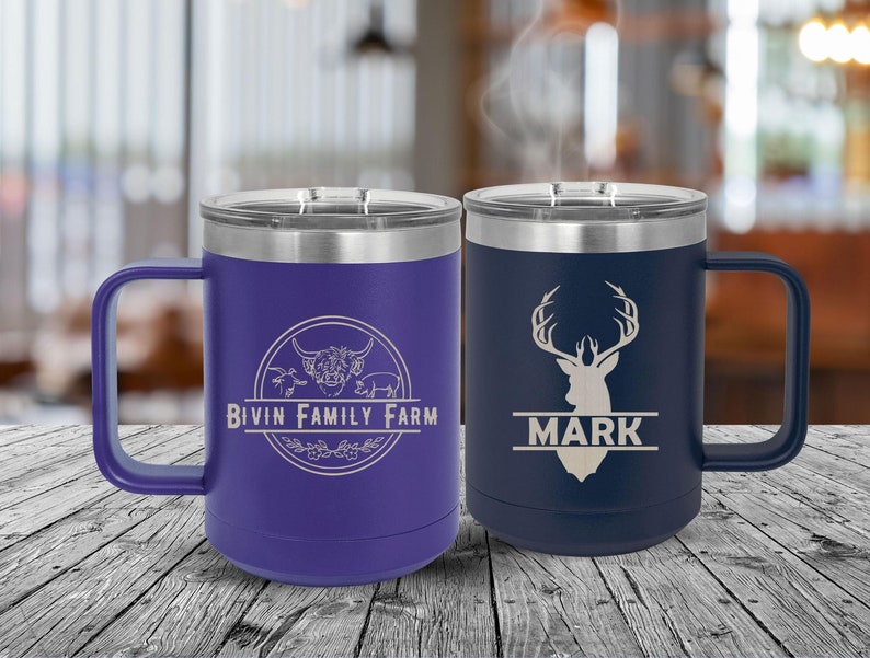May include: Two stainless steel travel mugs with handles. The mug on the left is purple with a white engraved logo that says "Bivin Family Farm" and a cow, pig, and sheep. The mug on the right is blue with a white engraved deer head and the name "Mark" underneath.
