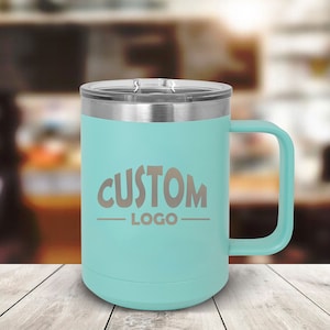 May include: A teal blue stainless steel travel mug with a lid and a handle. The mug has the words "CUSTOM LOGO" engraved on the side.