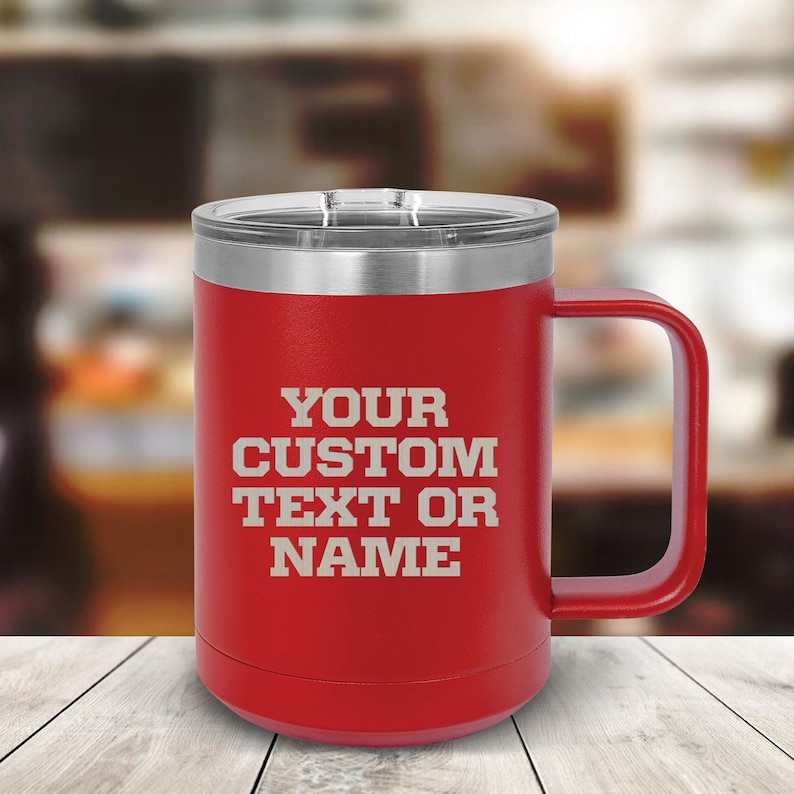 May include: Red stainless steel travel mug with a black handle. The mug has a white inscription that reads "YOUR CUSTOM TEXT OR NAME".