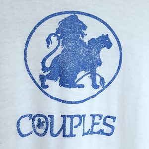 May include: White t-shirt with a blue graphic of a lion and a panther inside a circle. Below the graphic, the word "COUPLES" is printed in blue, with a small clover design. The shirt has a vintage, distressed look.