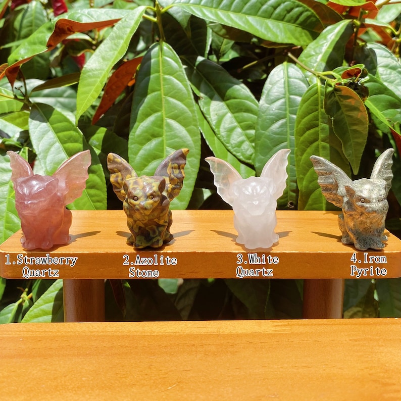 May include: Four carved gargoyle figurines made of different types of stone. The gargoyles are arranged in a row on a wooden shelf. From left to right, the gargoyles are made of strawberry quartz, azurite stone, white quartz, and iron pyrite.